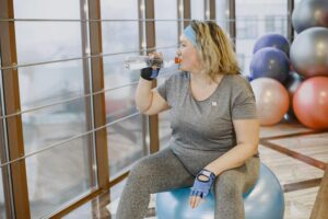 A woman drinks water while sitting on an exercise ball in a gym setting.