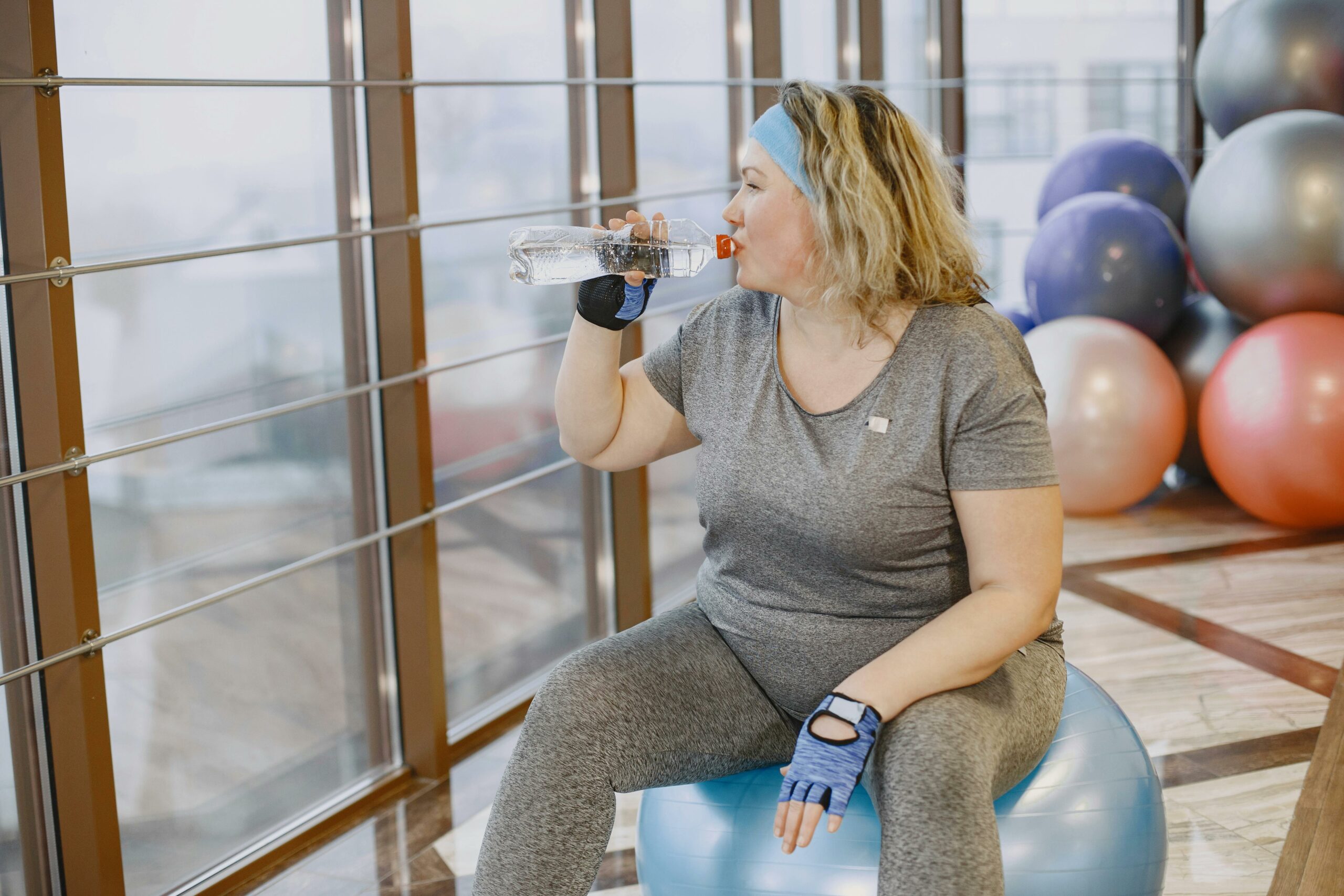 A woman drinks water while sitting on an exercise ball in a gym setting.